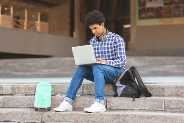 Young man typing on laptop outside university campus